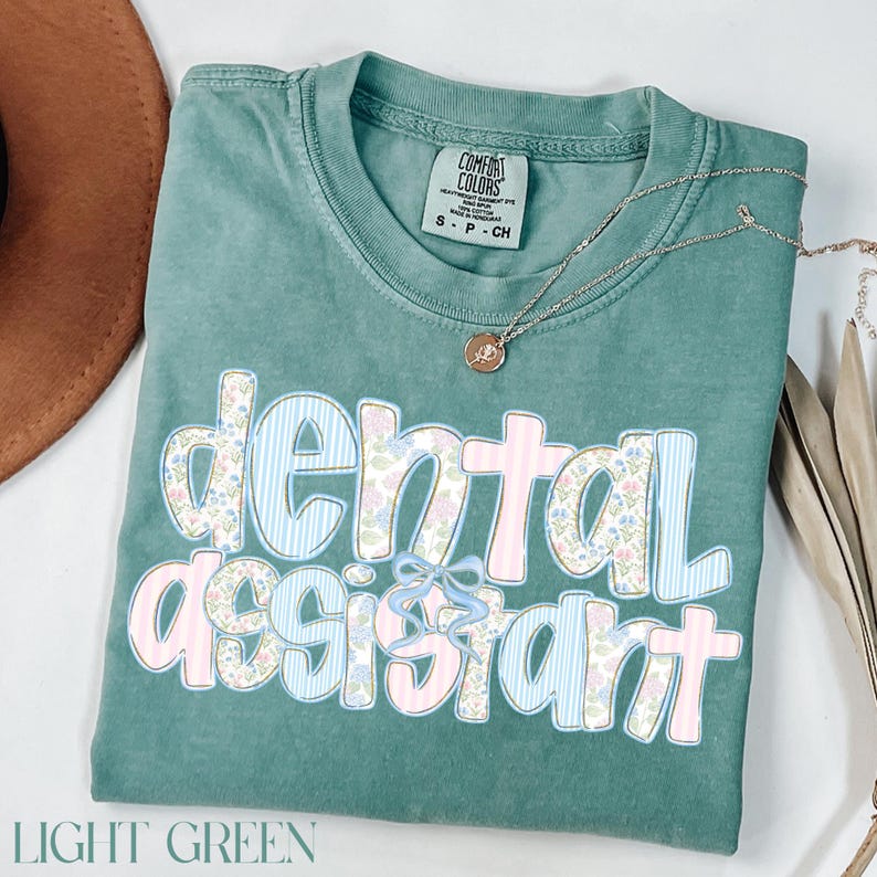 Op de afbeelding: Lichtgroen t-shirt met de woorden "dental assistant" in een bloemen- en streepjesontwerp. Het shirt heeft een ronde hals en korte mouwen. Een zilveren ketting met een hanger is over het shirt gedrapeerd.