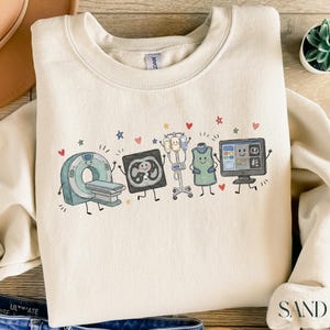 Peut inclure: Un sweat-shirt à col rond couleur sable orné d'une impression graphique d'équipements d'imagerie médicale anthropomorphes, incluant un scanner CT, une radiographie, un pied à perfusion, un tablier plombé et un écran, avec des cœurs et étoiles.