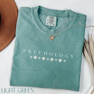 May include: Light green t-shirt with the text "Psychology" and a row of white daisy flowers. The shirt is made of a soft, comfortable fabric.