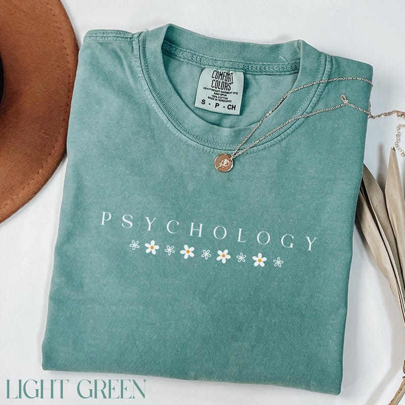 May include: Light green t-shirt with the text "Psychology" and a row of white daisy flowers. The shirt is made of a soft, comfortable fabric.