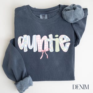 May include: Auntie sweatshirt in denim blue. The word "auntie" is in a bubble font with a patchwork design of blue stripes, pink gingham, floral patterns, and green gingham. A pink bow is below the word. The word "DENIM" is at the bottom right.