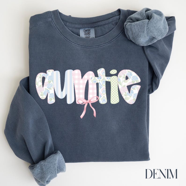 May include: Auntie sweatshirt in denim blue. The word "auntie" is in a bubble font with a patchwork design of blue stripes, pink gingham, floral patterns, and green gingham. A pink bow is below the word. The word "DENIM" is at the bottom right.