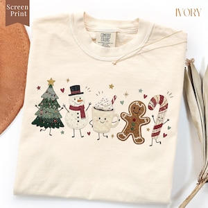 May include: An ivory t-shirt featuring a screen print graphic of five festive characters: a decorated Christmas tree, a snowman with a red scarf, a mug of hot cocoa with whipped cream, a gingerbread person, and a candy cane, all with faces.