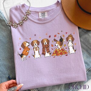 Puede incluir: Una camiseta de color morado claro con un gr&aacute;fico de cinco perros beagle con atuendos y poses oto&ntilde;ales, incluyendo uno con un su&eacute;ter de calabazas, uno con una taza de caf&eacute;, uno con una bufanda de cuadros, uno saltando en hojas y uno con gorro.