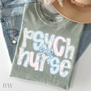 May include: Sage green t-shirt with the text "PSYCH NURSE" in a floral and striped design. The shirt is folded on top of blue jeans, with a gold necklace and a tan hat visible in the background.