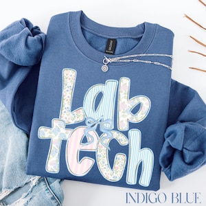 May include: Indigo blue sweatshirt featuring the text "Lab Tech" in a floral and striped pattern. A silver necklace with a small pendant is placed on top. The sweatshirt is folded on a white surface.