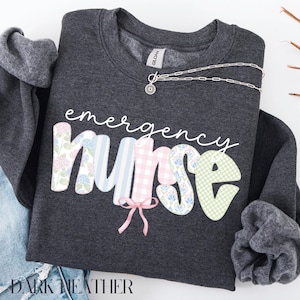 Puede incluir: Sudadera gris oscuro con las palabras "emergency nurse" en escritura blanca. La palabra "nurse" est&aacute; en patrones florales, a rayas y de cuadros. Un collar plateado con un colgante est&aacute; sobre la sudadera.