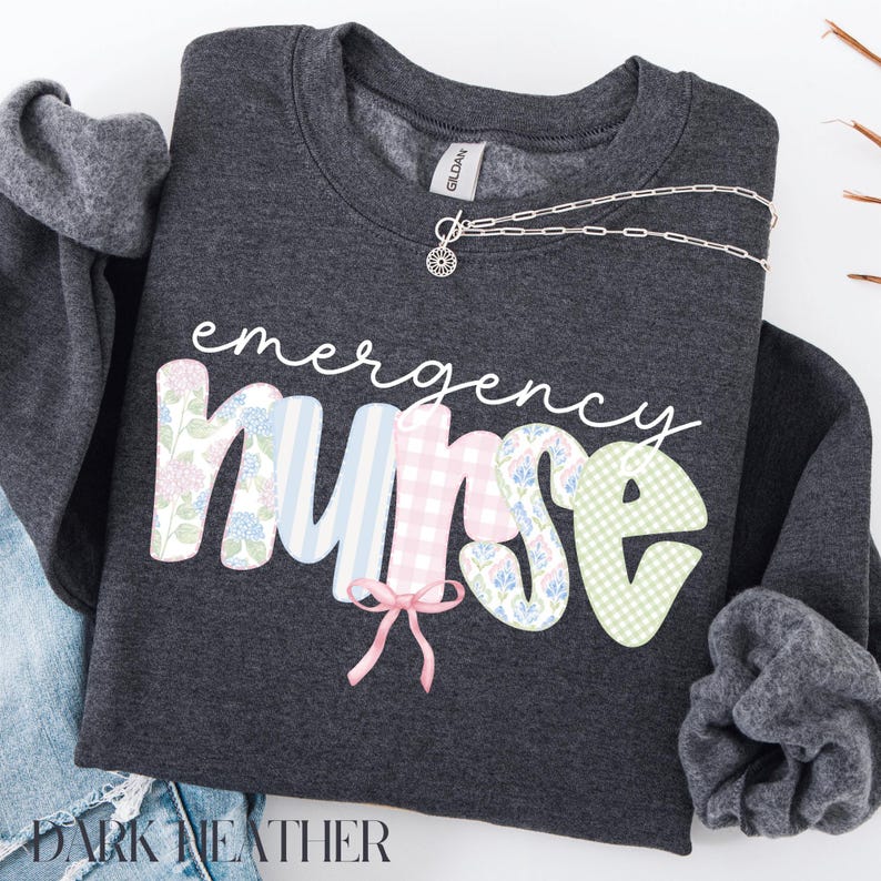 Puede incluir: Sudadera gris oscuro con las palabras "emergency nurse" en escritura blanca. La palabra "nurse" est&aacute; en patrones florales, a rayas y de cuadros. Un collar plateado con un colgante est&aacute; sobre la sudadera.
