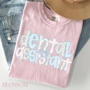 Op de afbeelding: Een lichtroze t-shirt met de tekst "dental assistant" in een bloemen- en streepjesontwerp. Het shirt ligt op een blauwe spijkerbroek, met een beige hoed en een ketting.