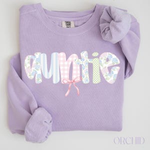 May include: Orchid-colored sweatshirt with the word "AUNTIE" in a whimsical font. The letters are decorated with floral and patterned fabrics, and a pink bow is tied beneath the word. A matching scrunchie rests on the shoulder.