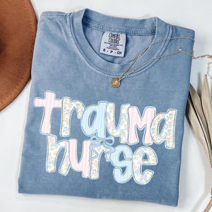 May include: A blue Comfort Colors t-shirt with the words "Trauma Nurse" in a floral and striped design. A gold necklace with a pendant is draped across the shirt. The shirt is folded and has a small tag.