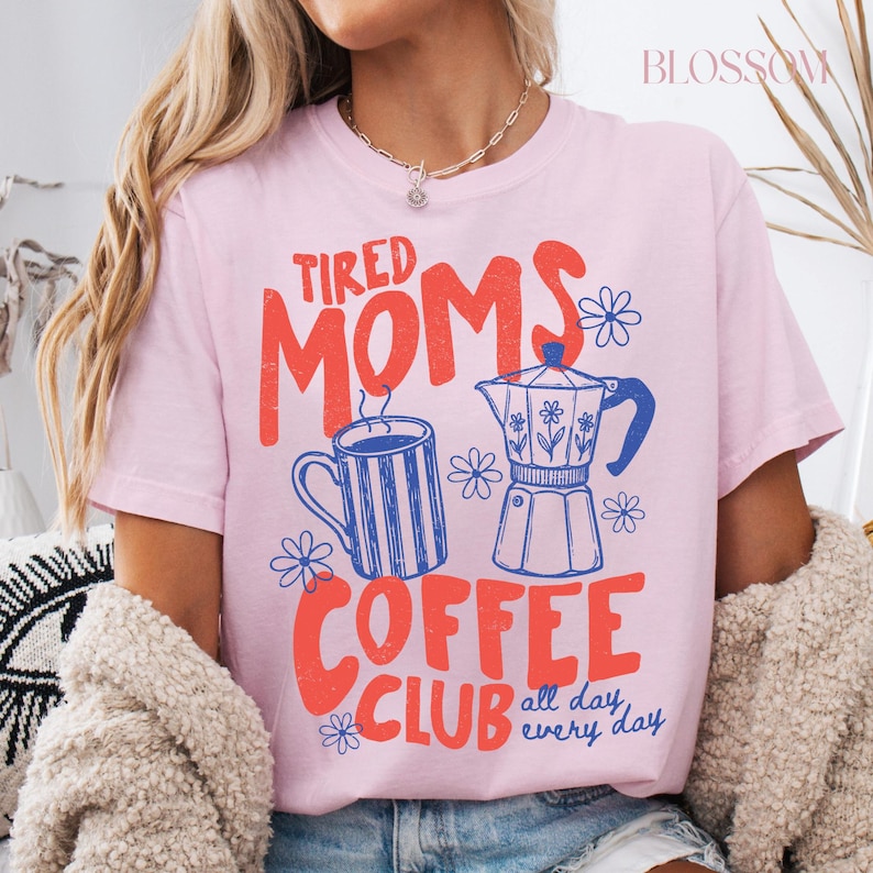 Puede incluir: Camiseta rosa claro con la frase "Tired Moms Coffee Club" en rojo y azul. El dise&ntilde;o incluye una taza de caf&eacute;, una cafetera y ilustraciones de flores. Tambi&eacute;n se incluye el texto "all day, every day".