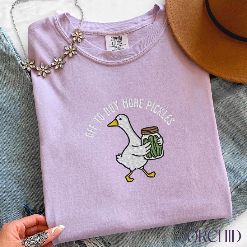 May include: A lilac Comfort Colors t-shirt featuring an embroidered cartoon goose carrying a jar of pickles. The text "OFF TO BUY MORE PICKLES" is arched above the goose. A brown hat and floral necklace are in the background.