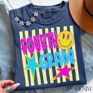 Op de afbeelding: Marineblauw T-shirt met de woorden "FOURTH GRADE" in roze en blauw, met een gele smiley en sterren. Het shirt heeft een ronde hals en is gemaakt van een zacht materiaal. Een ketting en een hoed staan ook op de foto.