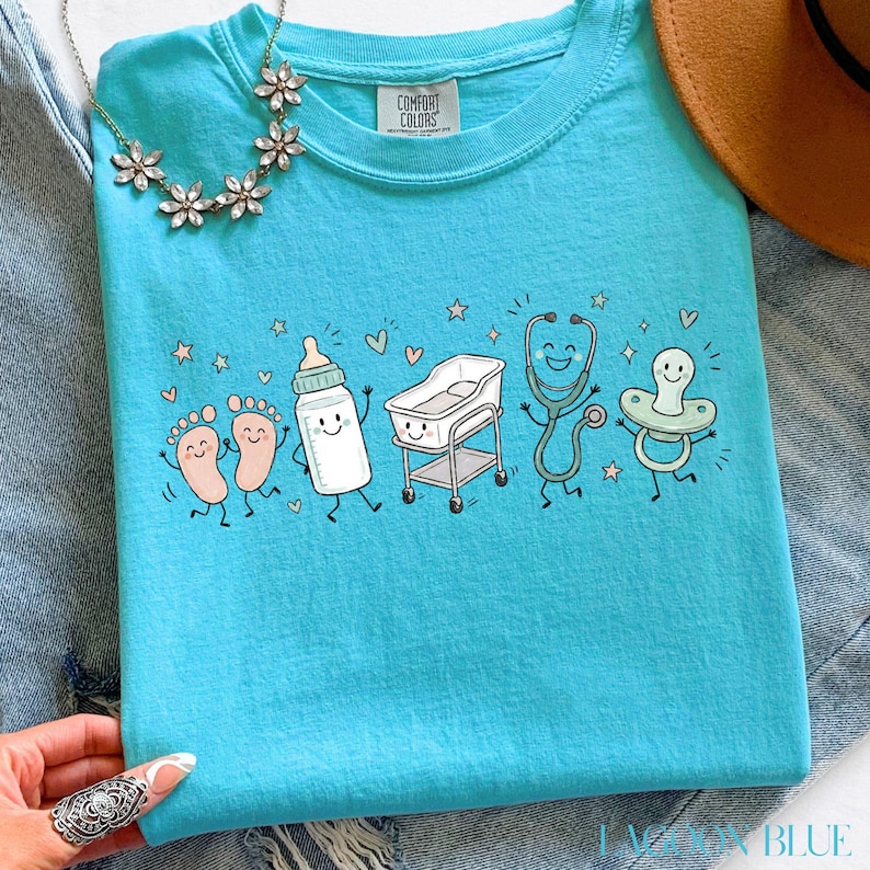 May include: A bright turquoise t-shirt featuring a graphic print of smiling baby-themed items including footprints, a baby bottle, a bassinet, a stethoscope, and a pacifier. The shirt has a crew neckline with a Comfort Colors brand label inside.