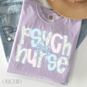 May include: A lilac t-shirt featuring the words "Psych Nurse" in a playful font. The letters are adorned with floral and striped designs, and a blue bow. The shirt is folded on top of a pair of blue jeans.