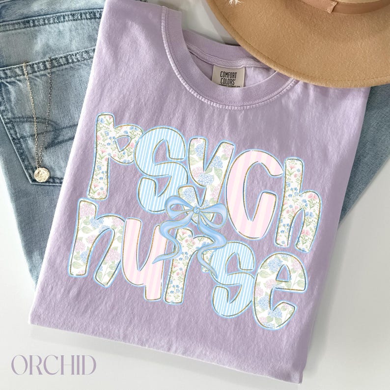 May include: A lilac t-shirt featuring the words "Psych Nurse" in a playful font. The letters are adorned with floral and striped designs, and a blue bow. The shirt is folded on top of a pair of blue jeans.