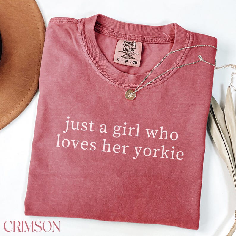 May include: Crimson-colored t-shirt with the text "just a girl who loves her yorkie" in white. A silver necklace with a round pendant rests on the shirt. The shirt has a crew neck and a tag that says "Comfort Colors".