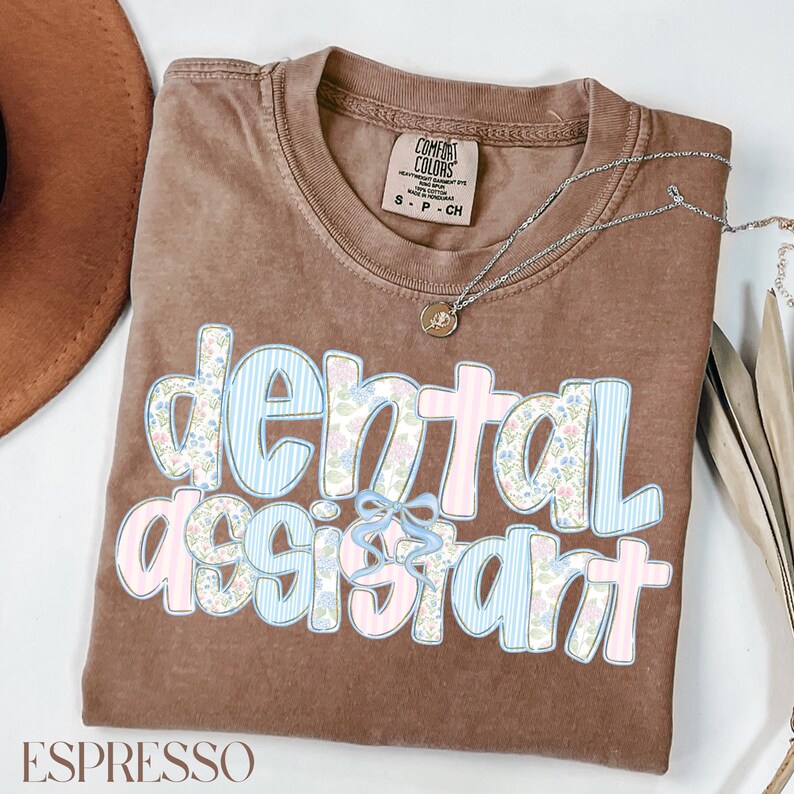 Op de afbeelding: Espresso-kleurig T-shirt met de tekst "dental assistant" in een bloemen- en streepjesontwerp. Het shirt heeft een ronde hals en korte mouwen. Een zilveren ketting met een kleine hanger is over het shirt gedrapeerd.