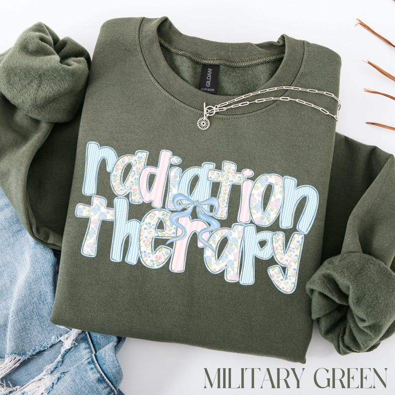 May include: A military green sweatshirt with the words "Radiation Therapy" in a floral and striped design. A silver necklace with a sunburst pendant is on the sweatshirt. The words "MILITARY GREEN" are at the bottom.