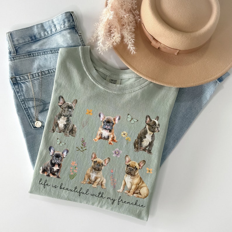May include: Sage green t-shirt featuring a graphic of six French Bulldogs and flowers. The text "life is beautiful with my frenchie" is printed on the shirt.