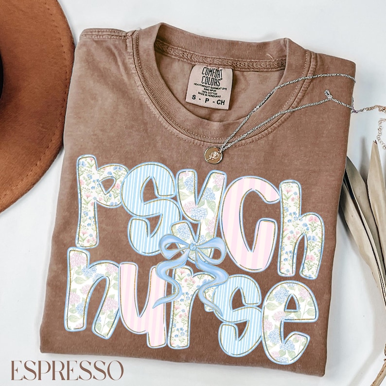 May include: Espresso-coloured t-shirt with the words "Psych Nurse" in a floral and striped design. A silver necklace with a pendant is draped across the shirt. The shirt is folded neatly.