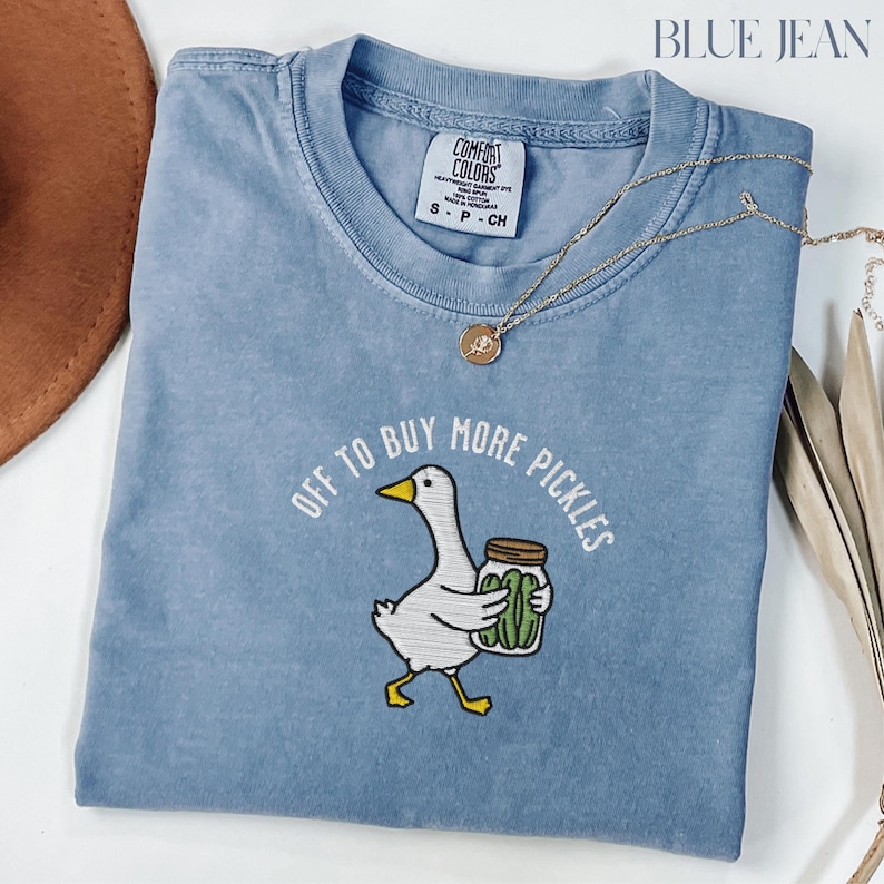 May include: A blue Comfort Colors t-shirt with the text "OFF TO BUY MORE PICKLES" and an embroidered graphic of a goose holding a jar of pickles. A gold necklace is draped across the shirt.
