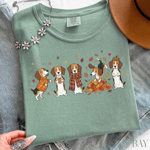 Puede incluir: Una camiseta verde salvia con un gr&aacute;fico de cinco perros beagle con ropa de oto&ntilde;o, incluyendo su&eacute;teres, bufandas y jugando entre hojas ca&iacute;das. La camiseta tiene un cuello redondo y una etiqueta de la marca Comfort Colors.