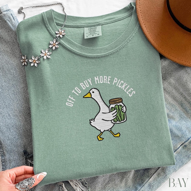 May include: A sage green t-shirt with the phrase "OFF TO BUY MORE PICKLES" and a cartoon goose carrying a jar of pickles. The shirt is folded with a necklace and a brown hat in the background.