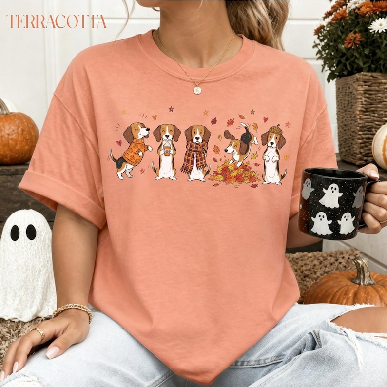 Puede incluir: Una camiseta de manga corta color melocot&oacute;n con un gr&aacute;fico de cinco perros beagle con ropa de oto&ntilde;o, incluyendo jers&eacute;is y bufandas, rodeados de hojas ca&iacute;das y corazones peque&ntilde;os. La palabra TERRACOTTA est&aacute; impresa en la esquina superior izquierda.