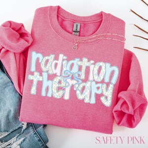 May include: A pink sweatshirt with the words "Radiation Therapy" in a floral and striped design. A light blue bow is between the words. A gold necklace with a pendant is on the shirt. The shirt is folded on top of blue jeans.