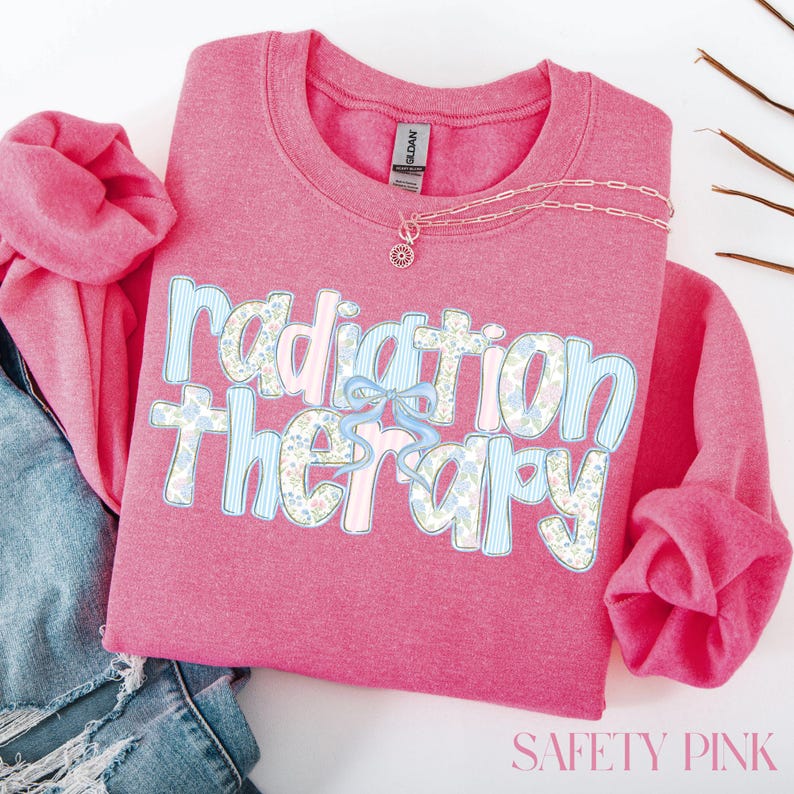 May include: A pink sweatshirt with the words "Radiation Therapy" in a floral and striped design. A light blue bow is between the words. A gold necklace with a pendant is on the shirt. The shirt is folded on top of blue jeans.