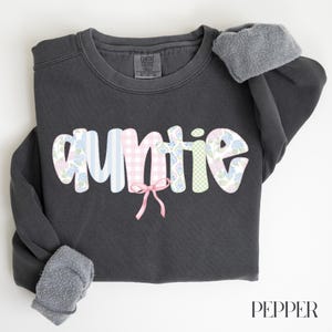 May include: A dark gray sweatshirt with the word "auntie" in a colorful, patterned design. The letters are decorated with blue stripes, pink gingham, floral patterns, and a pink bow. The sleeves have a gray, fuzzy lining.