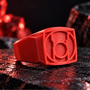 May include: A bright red ring featuring a square-shaped face with a circular emblem in the center. The ring has a textured surface with visible horizontal lines throughout its structure, indicating a 3D printed construction with a bold, solid design.
