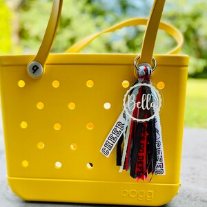 May include: A yellow tote bag with a black and white tassel keychain. The keychain has a white circle with the name "Bella" written in black. The tassel has black, white, red, and blue ribbons with the words "Cheer" and "Go Deep" written on them. The bag has the word "BOGG" written on the bottom.