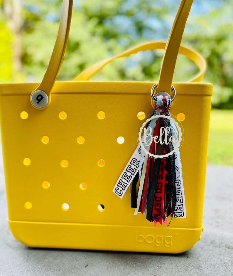 May include: A yellow tote bag with a black and white tassel keychain. The keychain has a white circle with the name "Bella" written in black. The tassel has black, white, red, and blue ribbons with the words "Cheer" and "Go Deep" written on them. The bag has the word "BOGG" written on the bottom.
