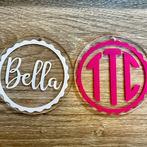 Op de afbeelding: Twee ronde acryl ornamenten met gegolfde randen. Een ornament is wit met de naam "Bella" geschreven in wit schrift. De andere ornament is roze met het monogram "TTC" in wit.