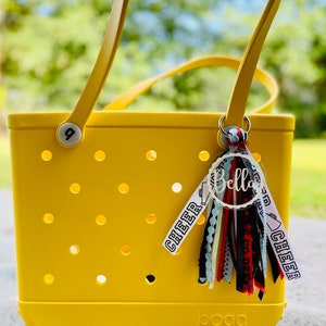 May include: Yellow tote bag with a white "bogg" logo and a clear round charm with the word "Bella" written in black. The charm is attached to a multi-colored tassel with black, red, white, and blue ribbons. The bag has a white tag with the word "cheer" written in black.