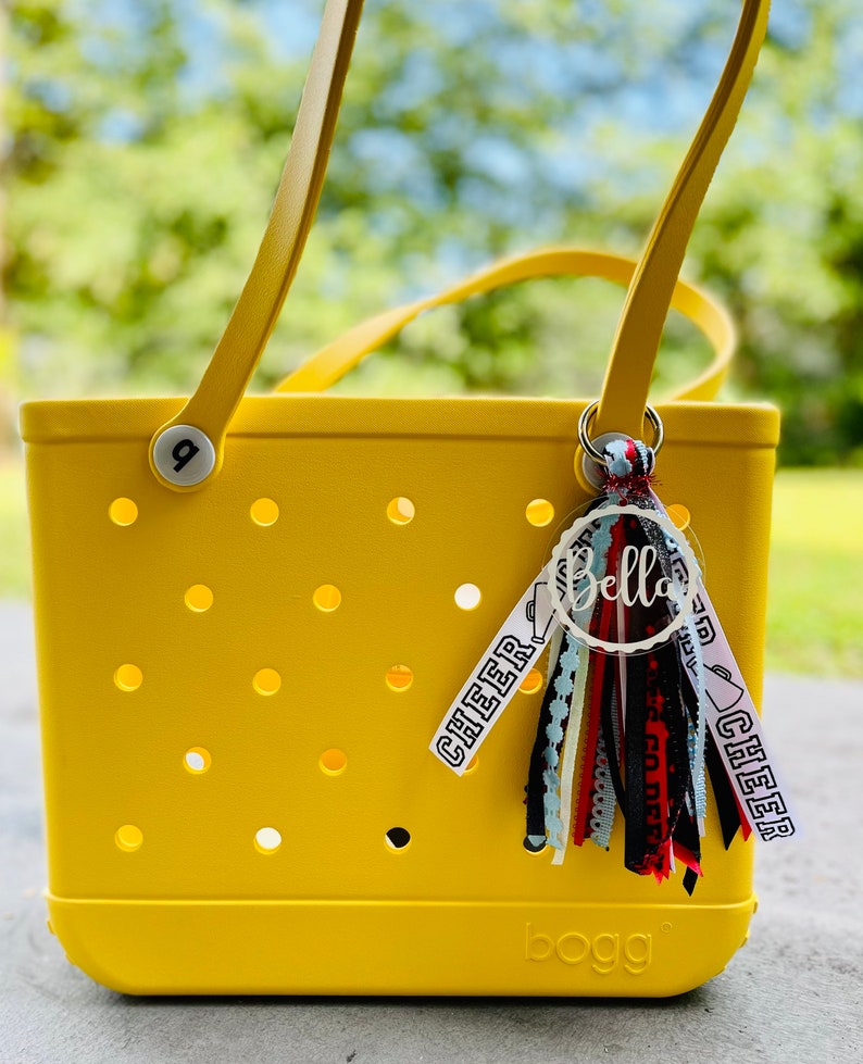 May include: Yellow tote bag with a white "bogg" logo and a clear round charm with the word "Bella" written in black. The charm is attached to a multi-colored tassel with black, red, white, and blue ribbons. The bag has a white tag with the word "cheer" written in black.