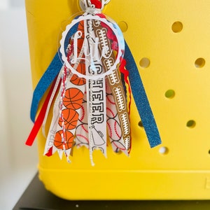May include: A yellow tote bag with a clear acrylic keychain with the name "Litte" written in white. The keychain is decorated with colorful ribbons featuring basketballs, baseballs, and footballs.
