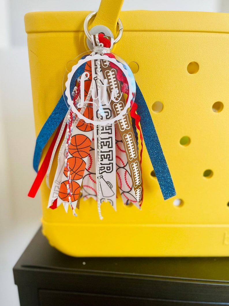 May include: A yellow tote bag with a clear acrylic keychain with the name "Litte" written in white. The keychain is decorated with colorful ribbons featuring basketballs, baseballs, and footballs.