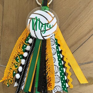 May include: A white volleyball keychain with the name "Mya" written in green on it. The keychain has a variety of ribbons in black, green, gold, and white with white pom-poms.