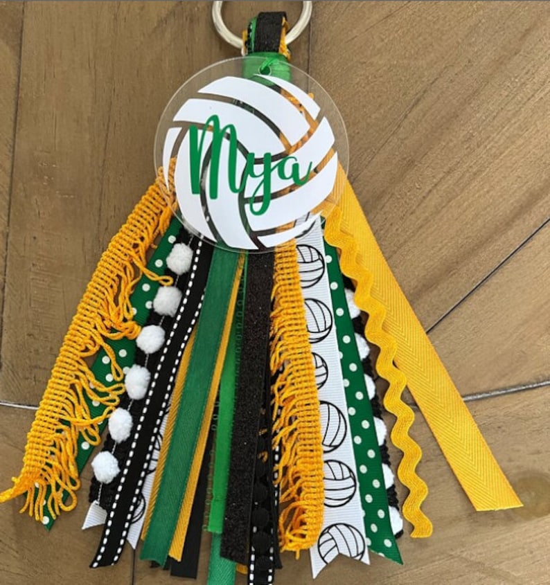 May include: A white volleyball keychain with the name "Mya" written in green on it. The keychain has a variety of ribbons in black, green, gold, and white with white pom-poms.