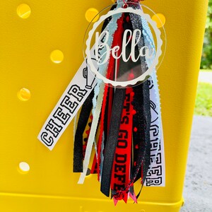 May include: A white acrylic charm with the name "Bella" in black script. The charm is attached to a black, red, and white tassel with the words "Cheer" and "Let's Go Defense" printed on the ribbons.