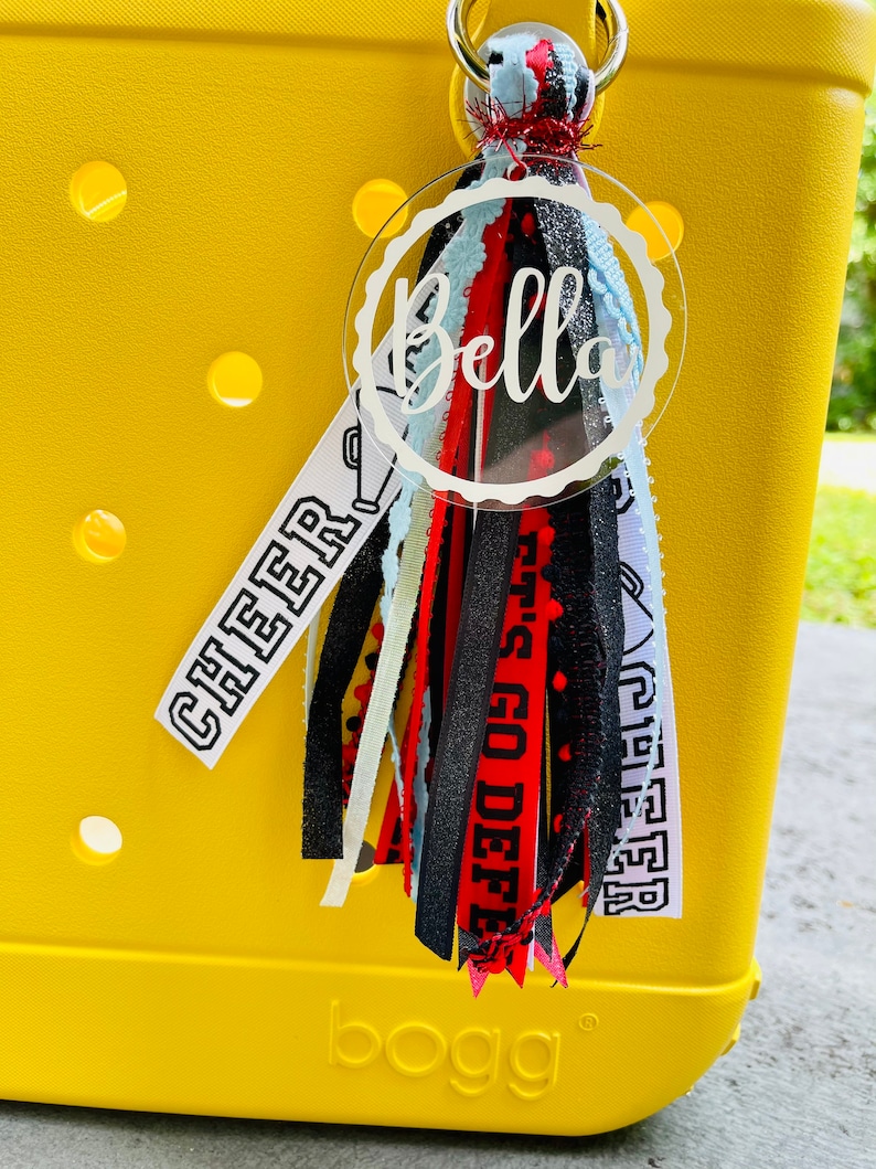 May include: A white acrylic charm with the name "Bella" in black script. The charm is attached to a black, red, and white tassel with the words "Cheer" and "Let's Go Defense" printed on the ribbons.