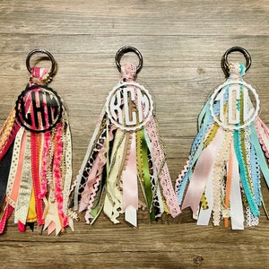 May include: Three keychains with different monogrammed initials, each with a circle of colorful ribbons attached. The initials are "KCM", "CHL", and "TEL".