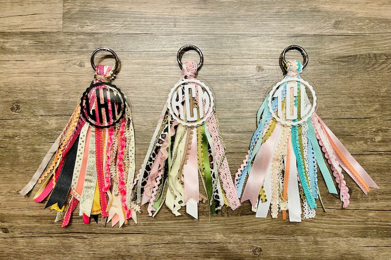 May include: Three keychains with different monogrammed initials, each with a circle of colorful ribbons attached. The initials are "KCM", "CHL", and "TEL".
