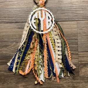 May include: A white acrylic keychain with the monogram "FYB" in a circle. The keychain has a gold metal ring and is adorned with a variety of colorful ribbons and tassels in shades of peach, navy blue, gold, and white.