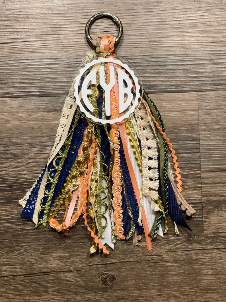 May include: A white acrylic keychain with the monogram "FYB" in a circle. The keychain has a gold metal ring and is adorned with a variety of colorful ribbons and tassels in shades of peach, navy blue, gold, and white.