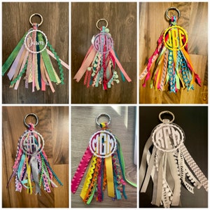 May include: Six keychains with colorful ribbons and a white circle with a monogram in the center. The monograms are "Dawn", "BPR", "JBF", "SAY", "MGM", and "RFM".